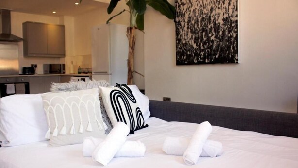 Una cama bien hecha con ropa de cama blanca y almohadones decorativos, una planta en una esquina y una cocina moderna al fondo.