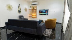 A modern living room with a dark sofa, a small armchair, and a TV mounted on the wall. - Peaceful Escape in Tema Community 25 (Dawhenya)