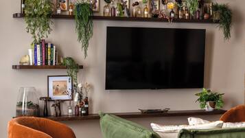 A living room with a TV, green and orange sofas, and shelves with plants and books.