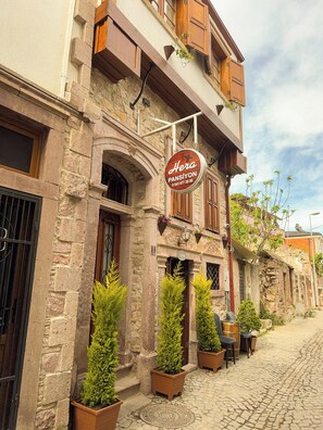 A stone building with a sign for "Hera Pansiyon" and a small outdoor seating area with potted plants. - Hera Pansiyon (Ayvalik)