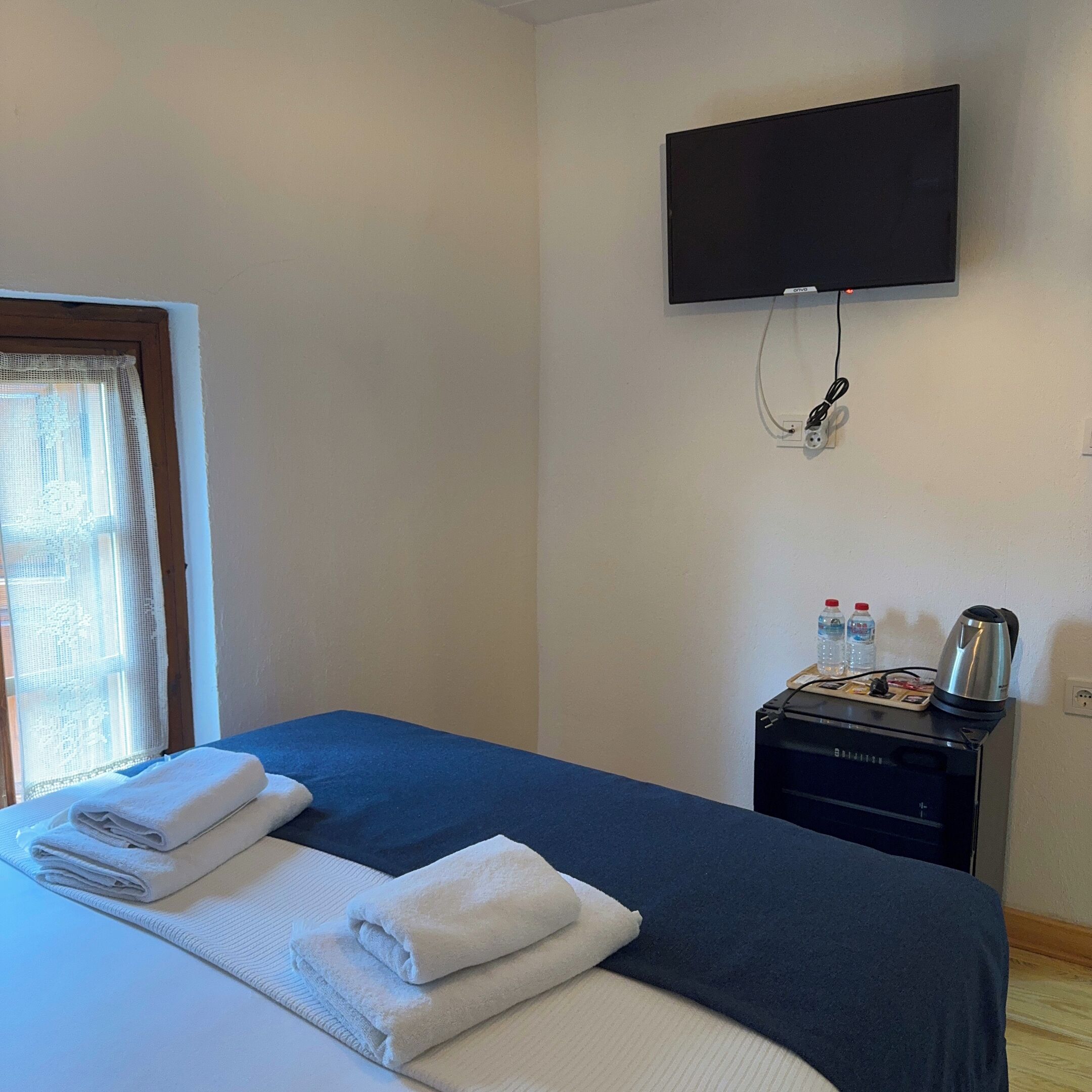 A hotel room with a bed, a television mounted on the wall, a small table with a kettle, and a window with a curtain.