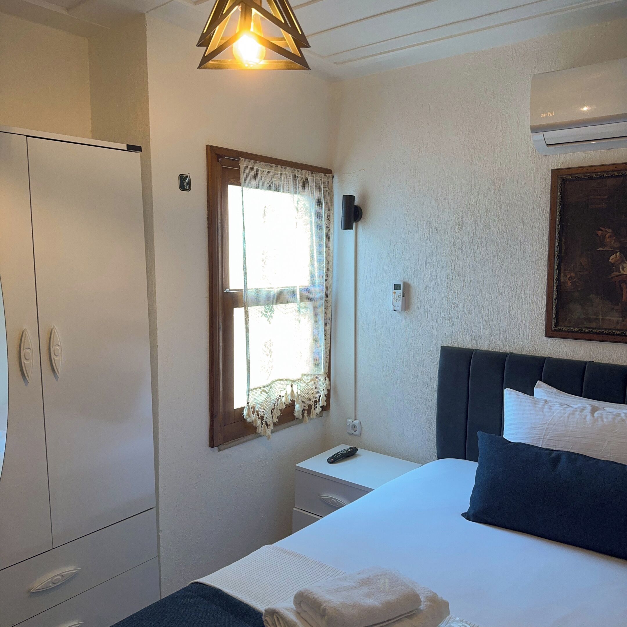 A bedroom with a bed, a wardrobe, a window with curtains, a bedside table, a lamp, and an air conditioning unit.
