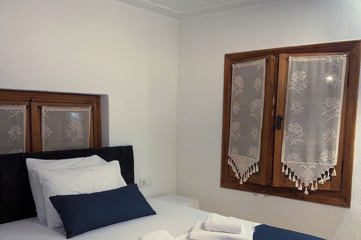 A neatly made bed with white and blue bedding, a wooden headboard, and a window with lace curtains.