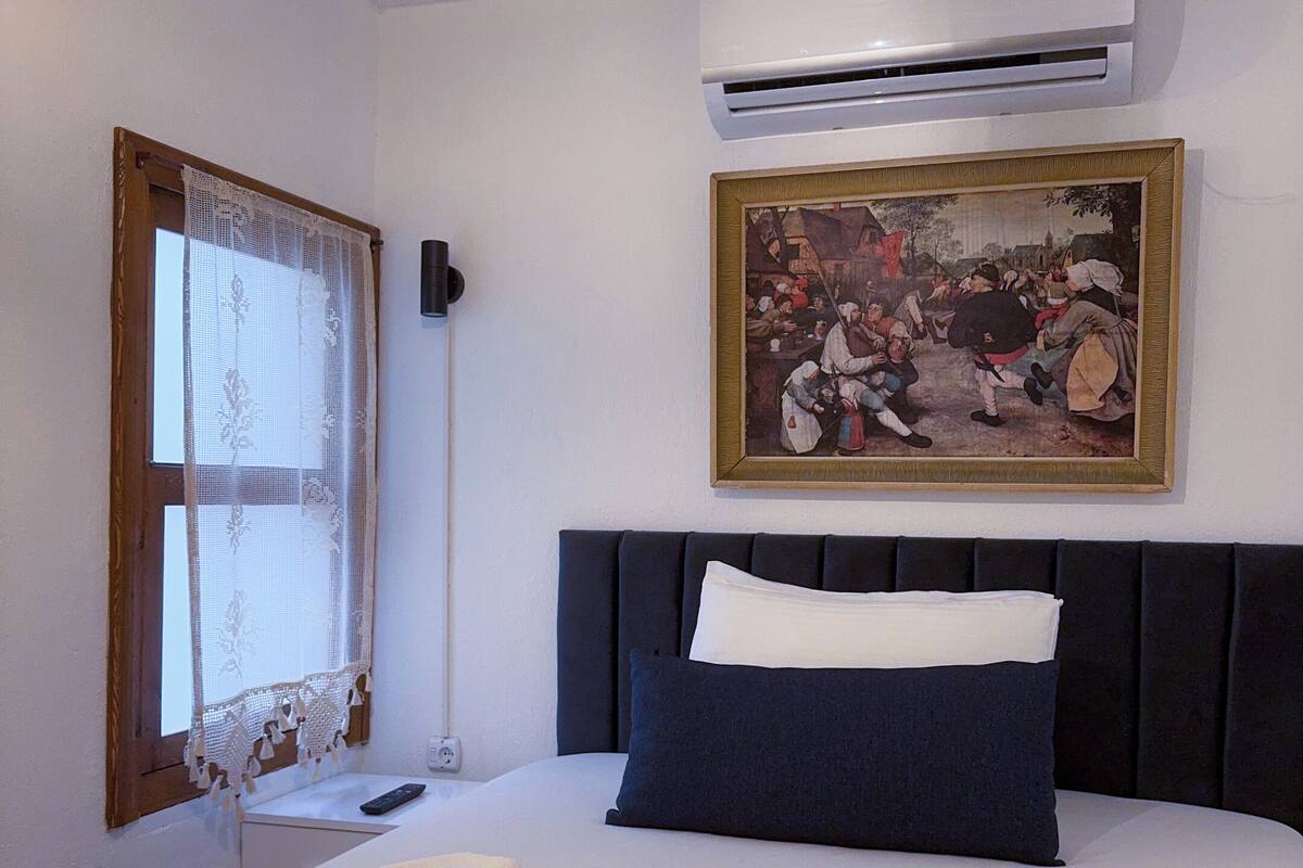 A hotel room with a bed, a painting, an air conditioner, and a window with lace curtains.