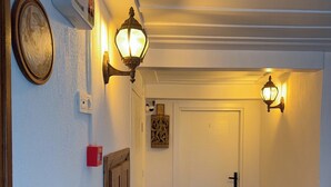 A hallway with a door, wall-mounted lights, a fire extinguisher, and a patterned rug. - Hera Pansiyon (Ayvalik)