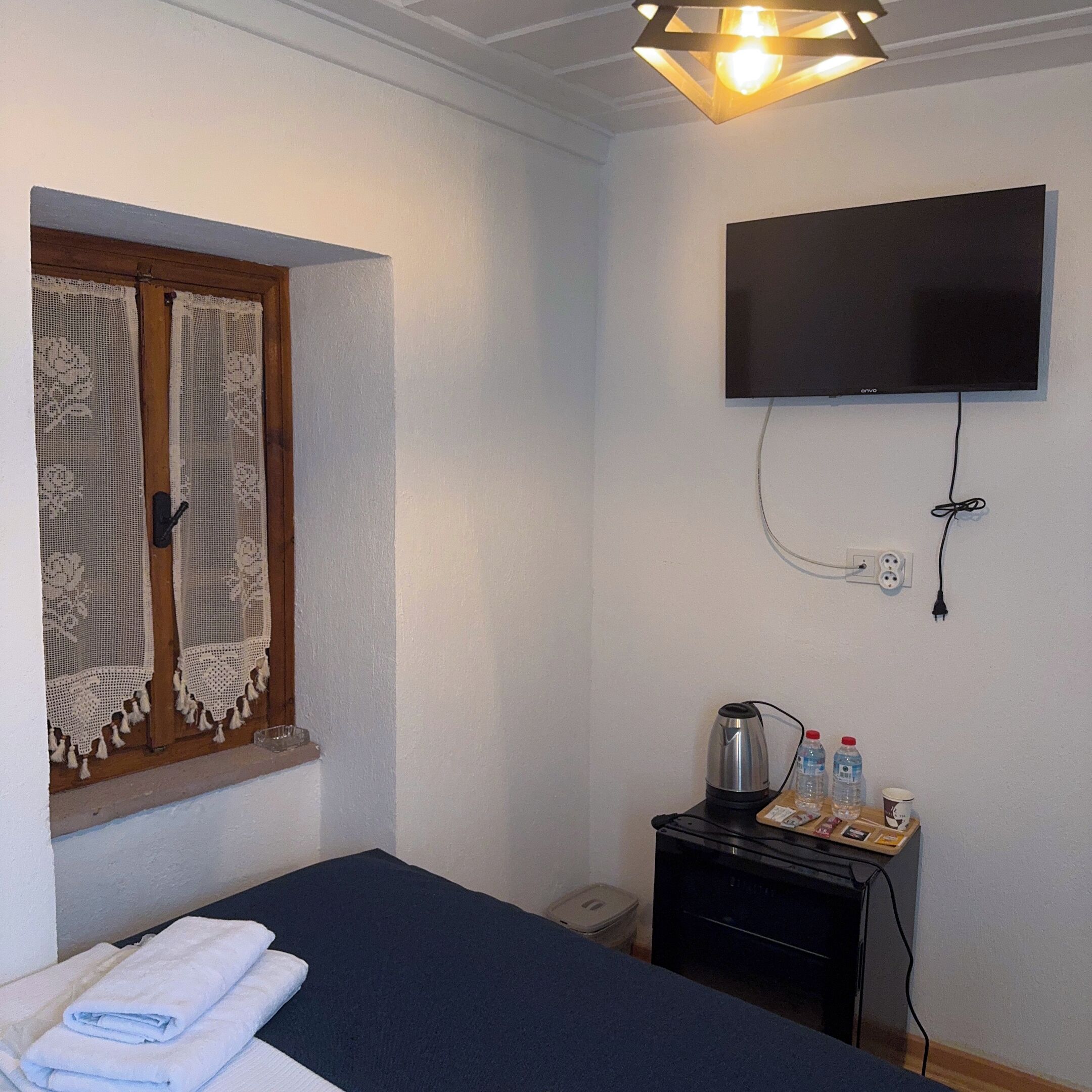 A room with a bed, a flat-screen TV mounted on the wall, a small table with a kettle and water bottles, and a window with lace curtains.