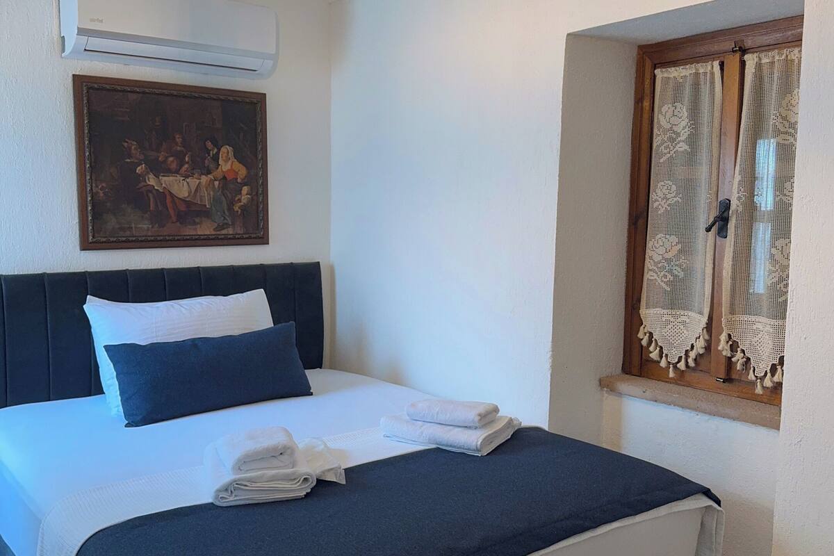 A neatly made bed with white and navy bedding, a painting above the bed, and a window with lace curtains.