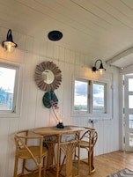 Um quarto com paredes brancas com painéis, uma mesa redonda com quatro cadeiras de vime, um espelho com moldura em forma de sol e duas luminárias fixadas na parede.