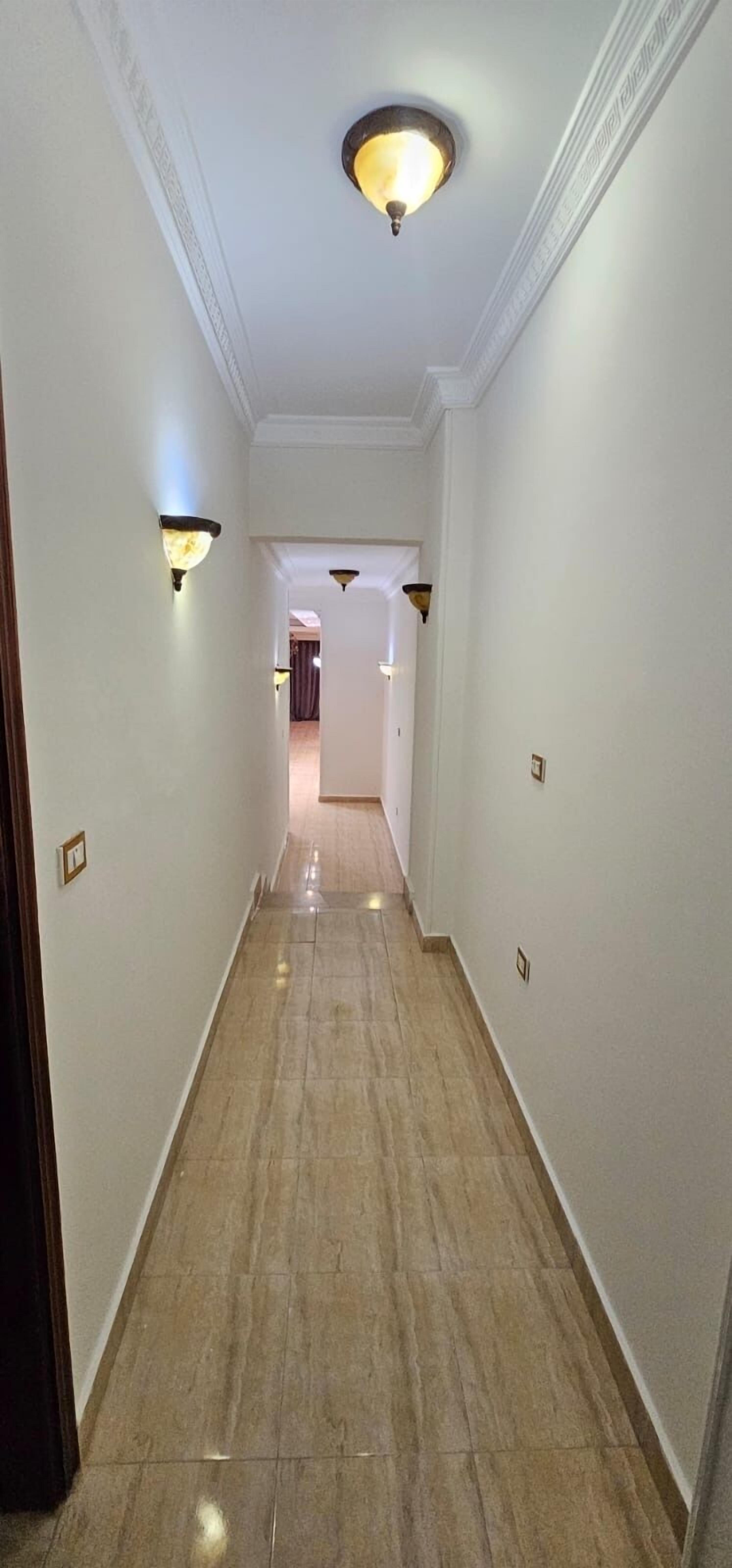 A hallway with marble flooring, white walls, and wall-mounted lighting fixtures.