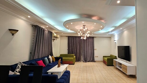 A modern living room with a blue sofa, green armchairs, a white TV stand, and a chandelier.