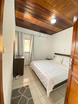 A bedroom with a wooden ceiling, a bed with white bedding, a nightstand, a window with curtains, and a small rug on the floor. - Chambres Bohèmes (Kribi)