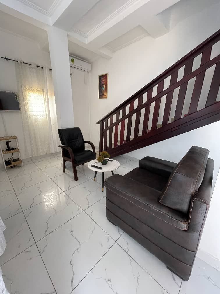 Una sala de estar con un sofá marrón oscuro, una silla negra, una mesita redonda con un florero y una escalera con baranda de madera.