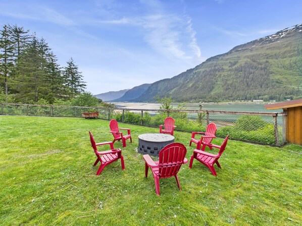 A patio with red Adirondack chairs and a round table, overlooking a lake and mountains. - Oceanfront Fire Pit, Decks, Large Yard, Views! (Juneau)
