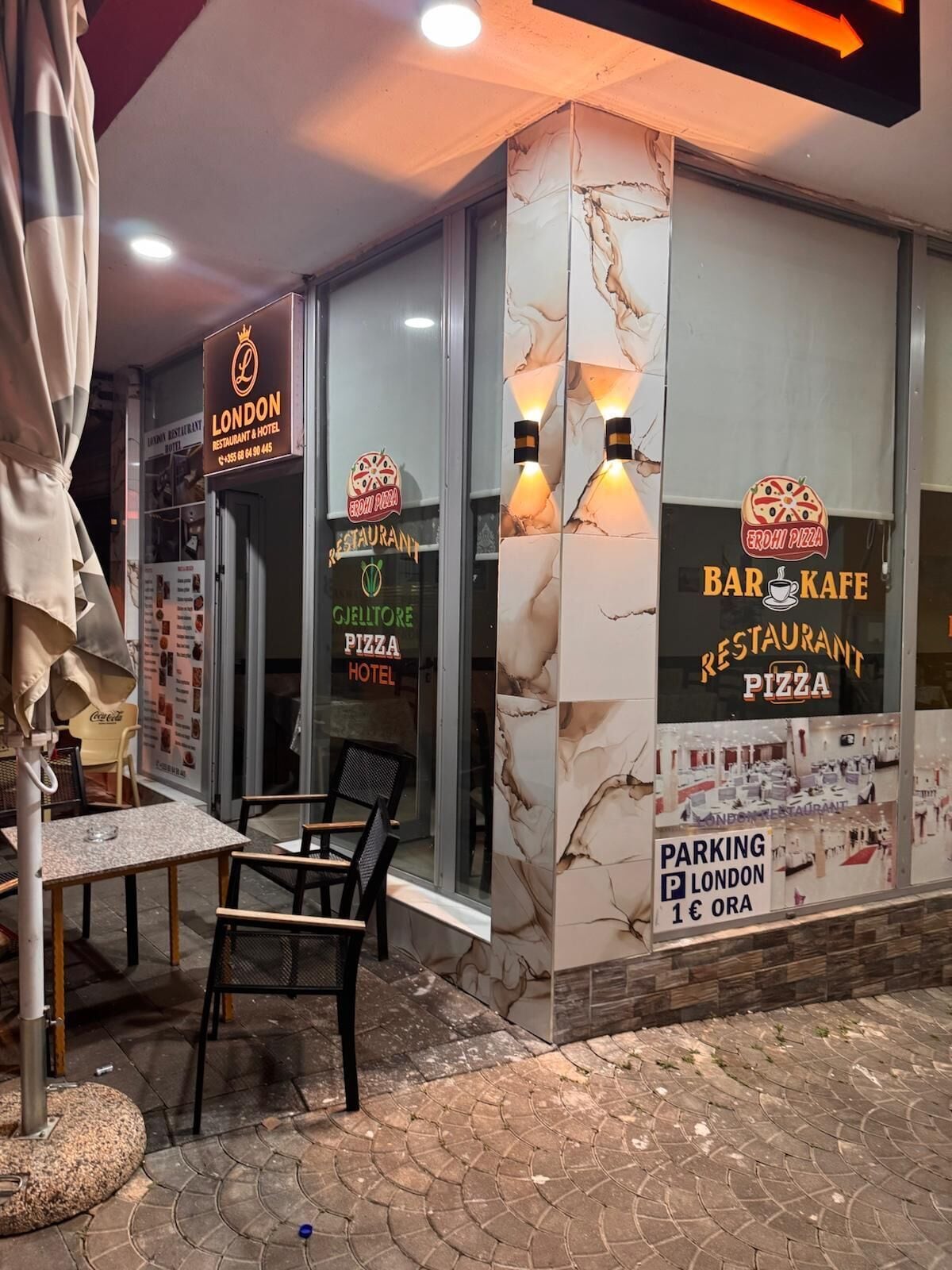 An exterior of a restaurant with a sign for 'London Restaurant & Hotel' and 'Gjelmire Pizza Hotel.' There are outdoor seating chairs and a table.