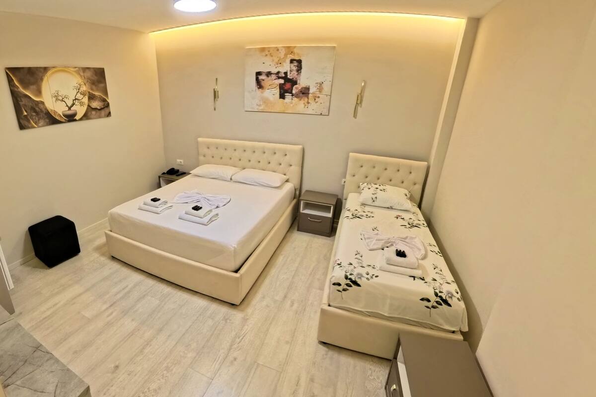 Un dormitorio con una cama grande, una cama más pequeña, una mesita de noche, un taburete negro y obras de arte en las paredes.