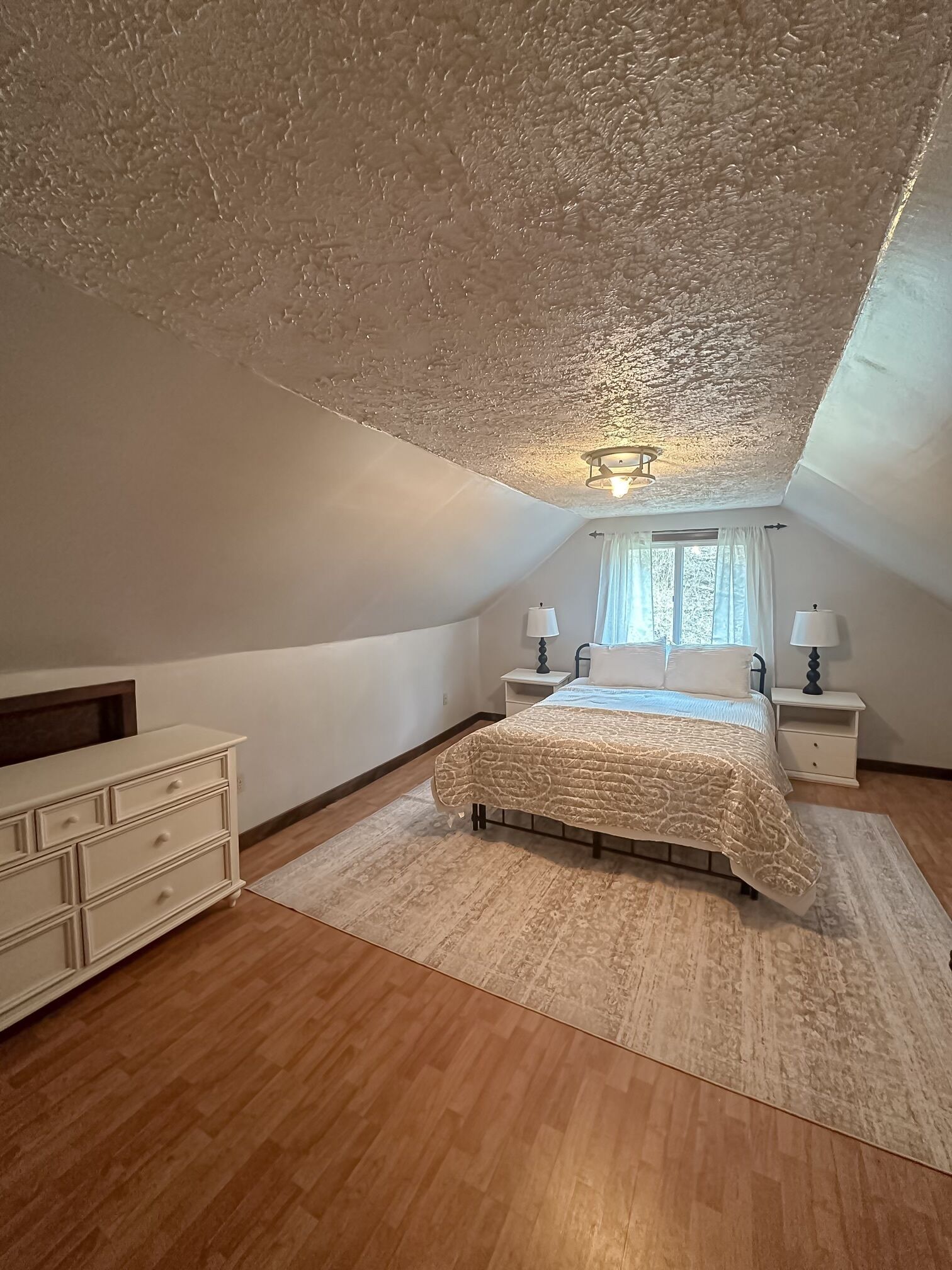 Une chambre avec un plafond en pente, un lit recouvert d’une couvre-lit à motifs, deux tables de chevet avec des lampes, une commode et une fenêtre avec des rideaux.
