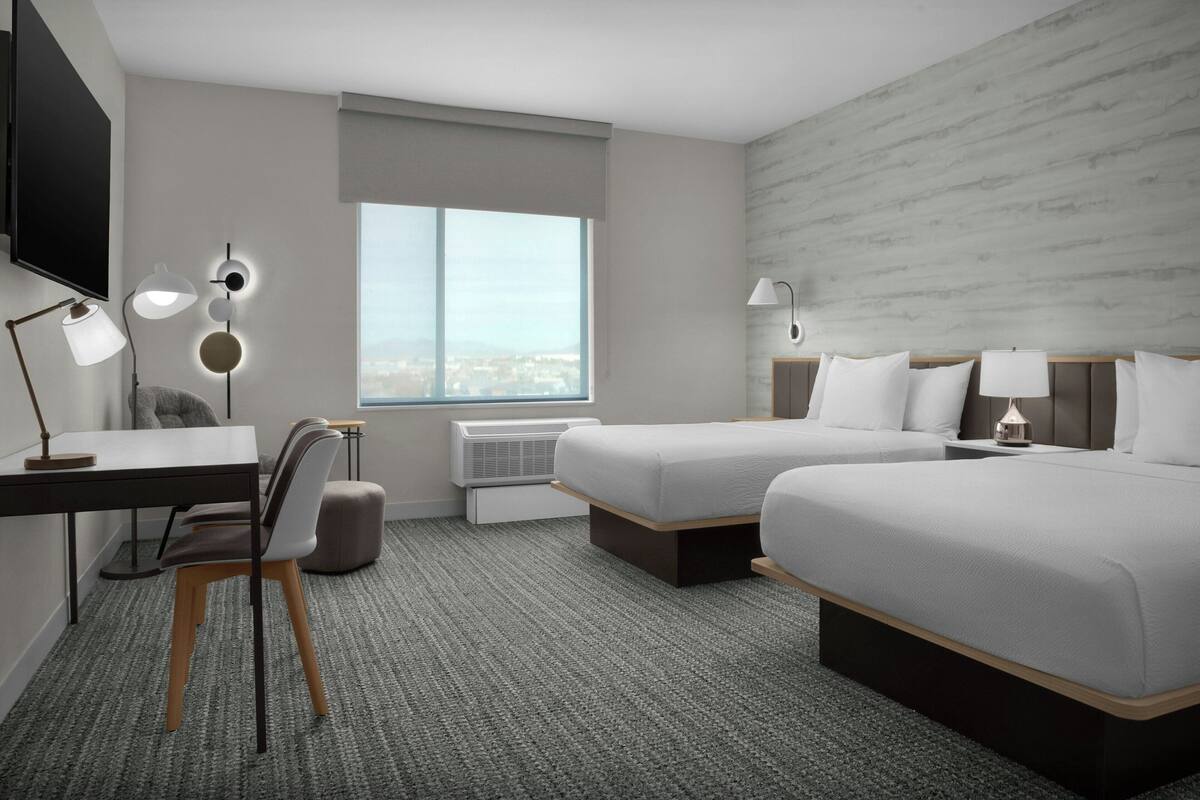 A hotel room with two beds, a desk, a chair, a TV, and a large window.