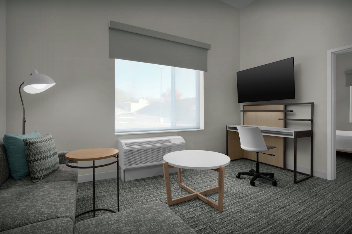 A modern room with a sofa, a round table, a desk with a chair, a flat-screen TV, and a large window.