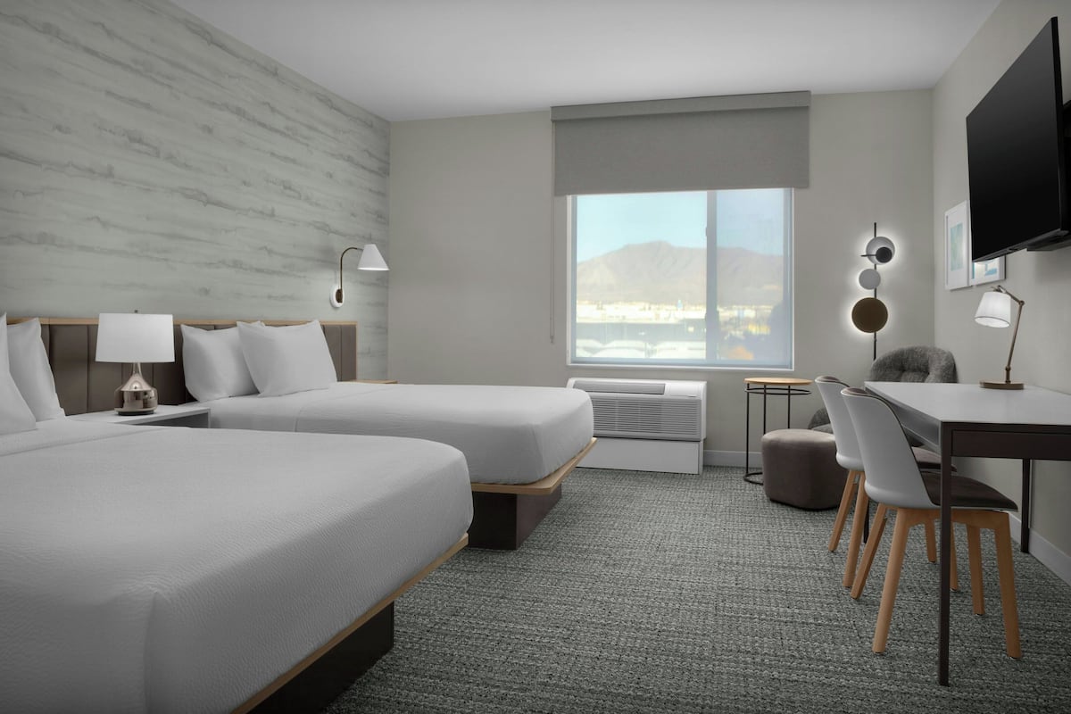 A hotel room with two beds, a desk, chairs, a TV, and a large window with a view.