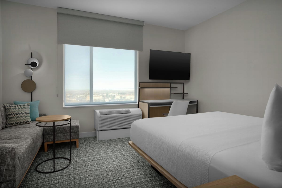 A hotel room with a bed, a desk, a chair, a small table, a sofa, a TV, and a large window.