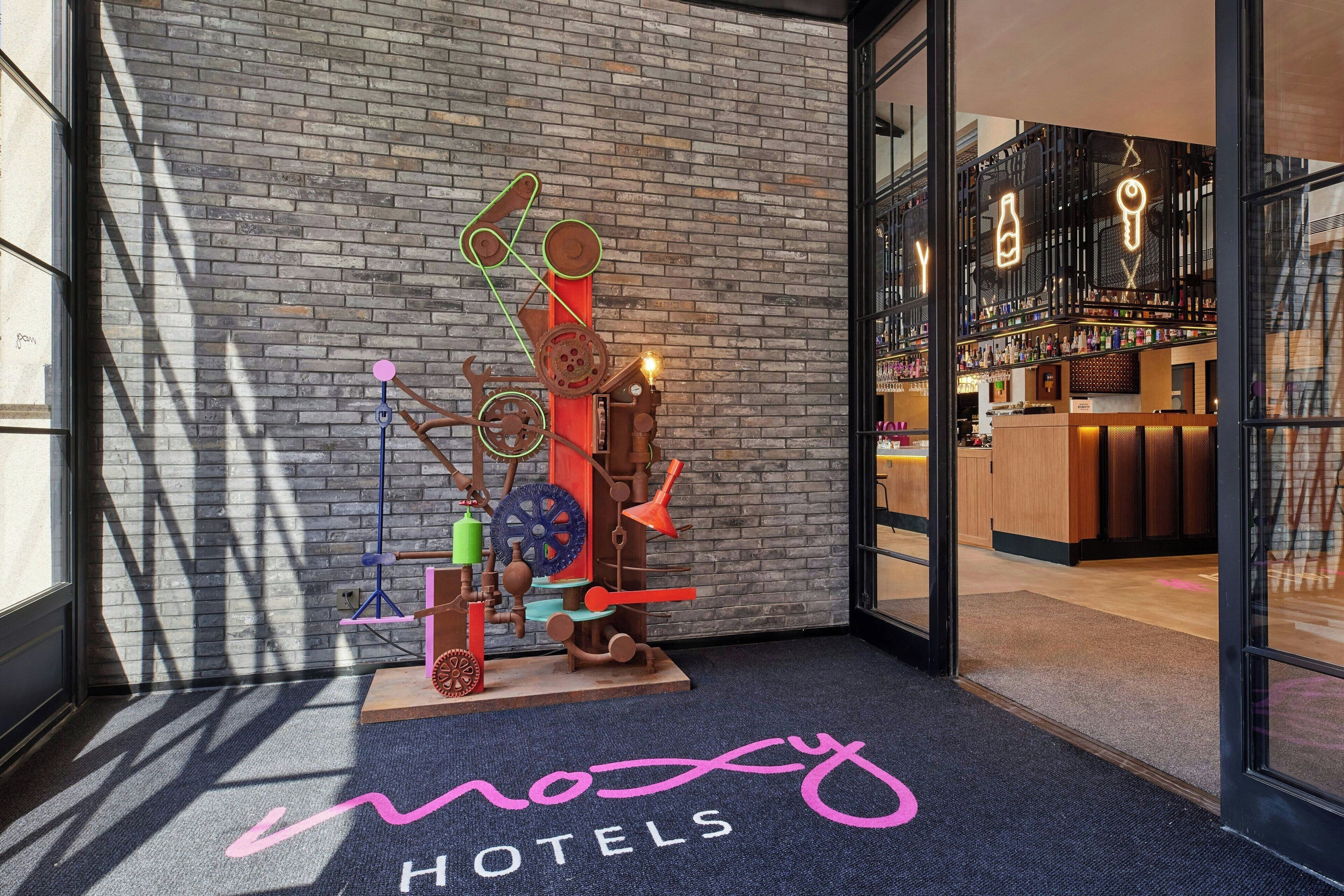 Entrada de hotel con una escultura abstracta colorida, zona de recepción con bar y el nombre del hotel en la alfombra.