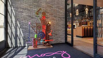 Entrada de hotel con una escultura abstracta colorida, zona de recepción con bar y el nombre del hotel en la alfombra.
