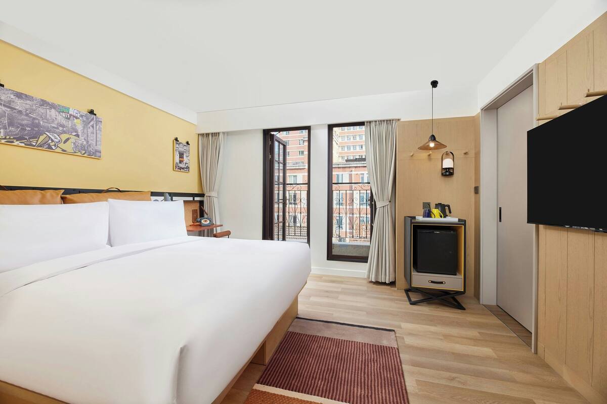 A hotel room with a large bed, a TV, and a balcony with a view of buildings.
