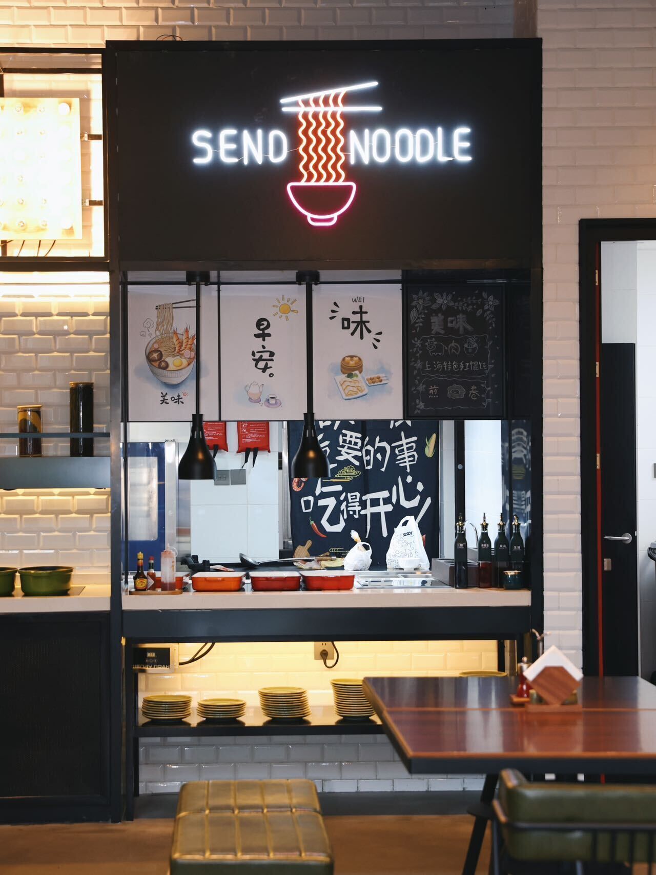 En restauranginteriör med en neonreklamskylt som visar texten ”SEND NOODLE”, en disk med olika rätter och tillbehör samt ett område med bord och stolar där gästerna kan sitta.