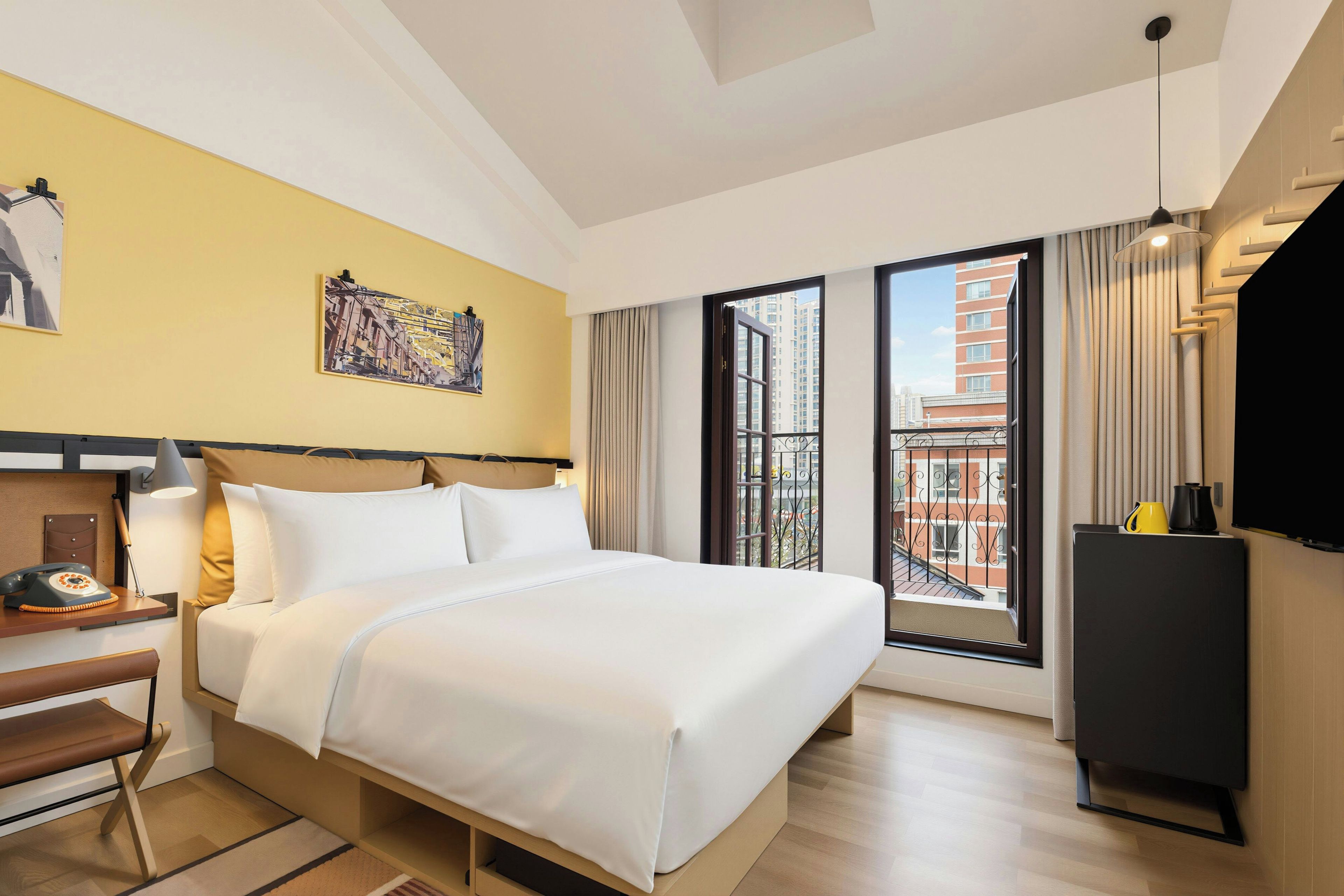 A hotel room with a bed, a desk with a telephone, a painting on the wall, and a window with a view of buildings.