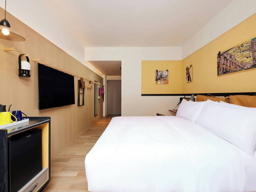 A hotel room with a bed, a flat-screen TV, a minibar, and artwork on the walls.