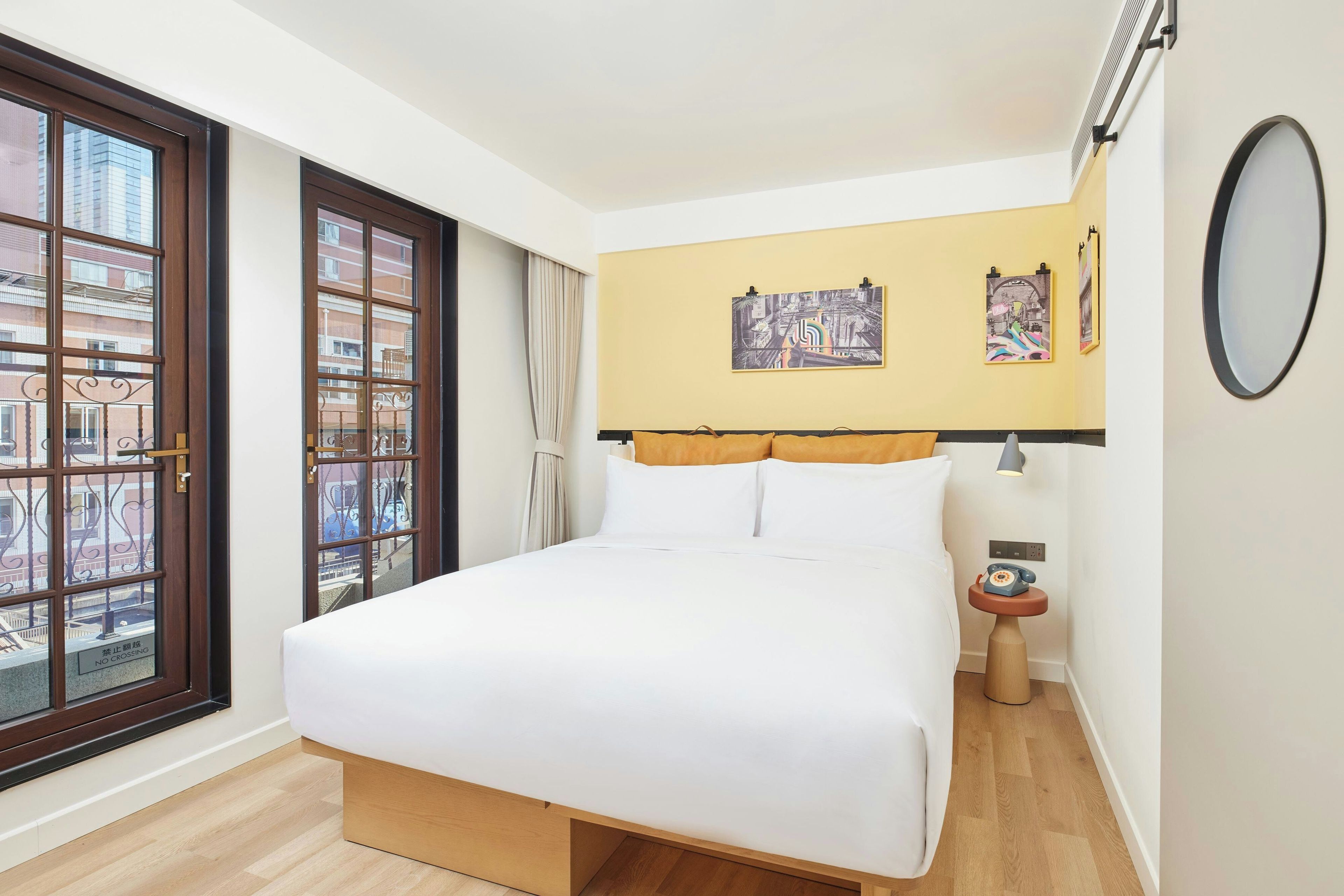 Habitación de hotel con un ventanal grande, una cama con ropa blanca, una mesita de noche con lámpara y dos obras de arte colgadas en la pared.