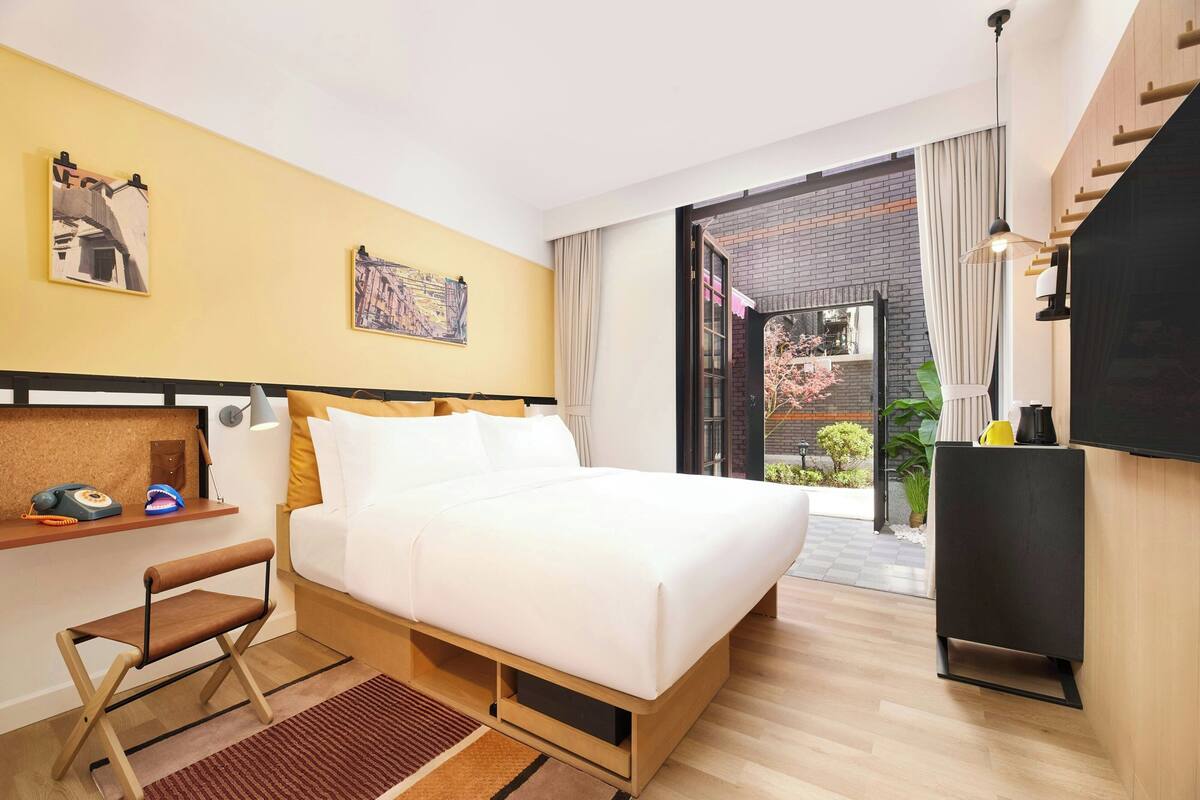 A modern hotel room with a large bed, a desk with a chair, a TV, and a view of the outside.