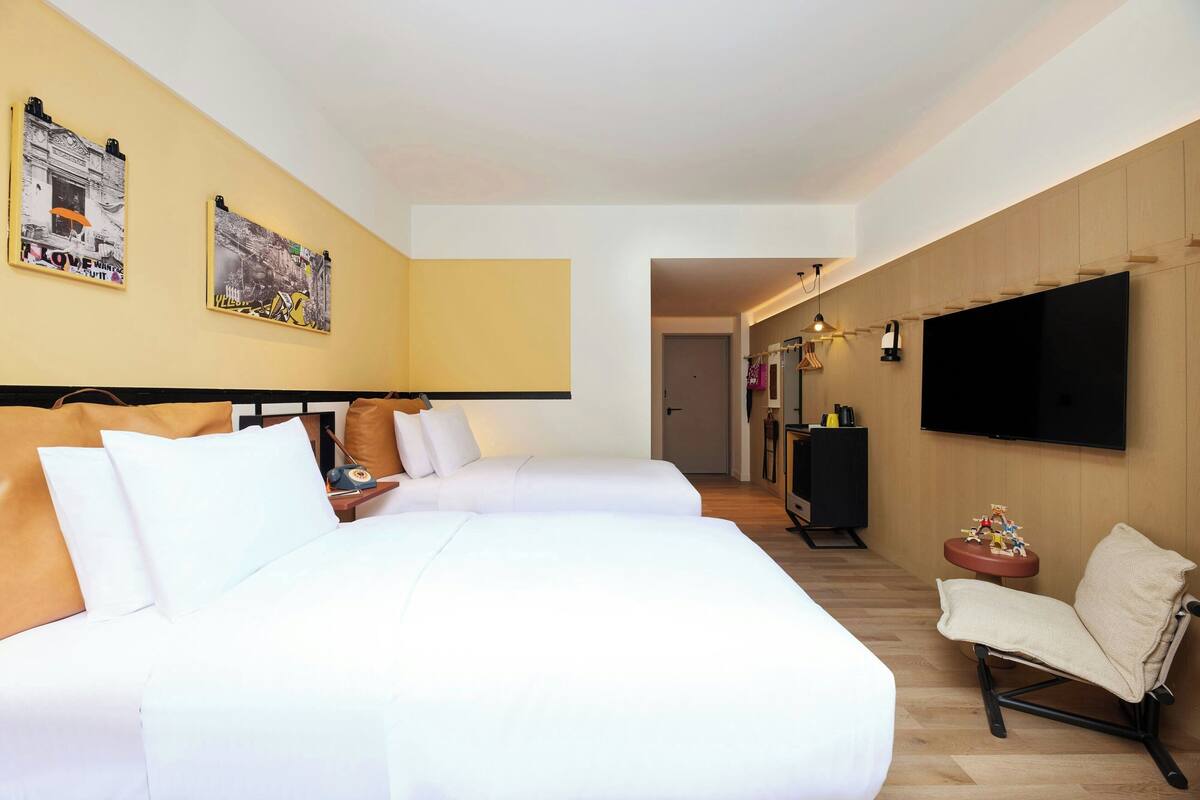 A hotel room with two beds, a TV, a small table with a lamp, and a chair.