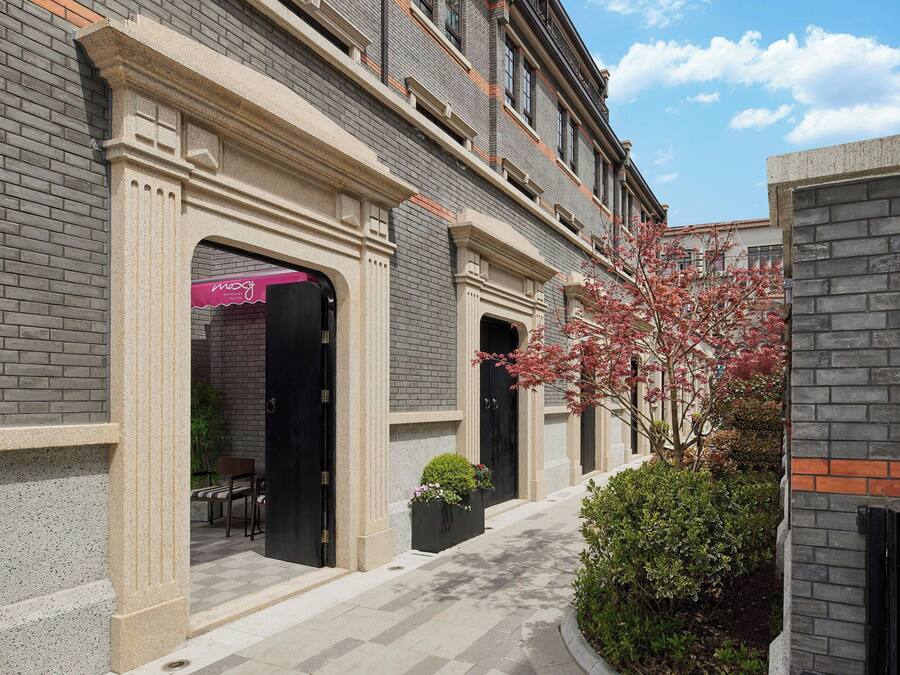 Un edificio con fachada de ladrillo, una entrada central con un arco decorativo y un pequeño jardín con un árbol y arbustos.