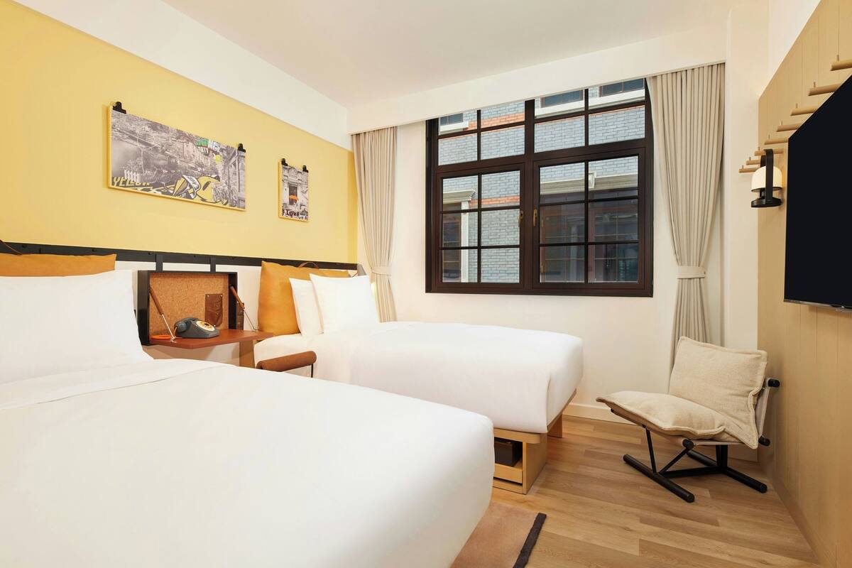 A hotel room with two beds, a chair, a desk with a telephone, and a large window with curtains.