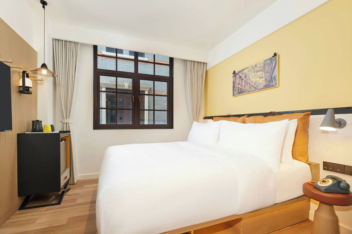 A hotel room with a large bed, a desk with a lamp, a telephone, and a window with curtains.