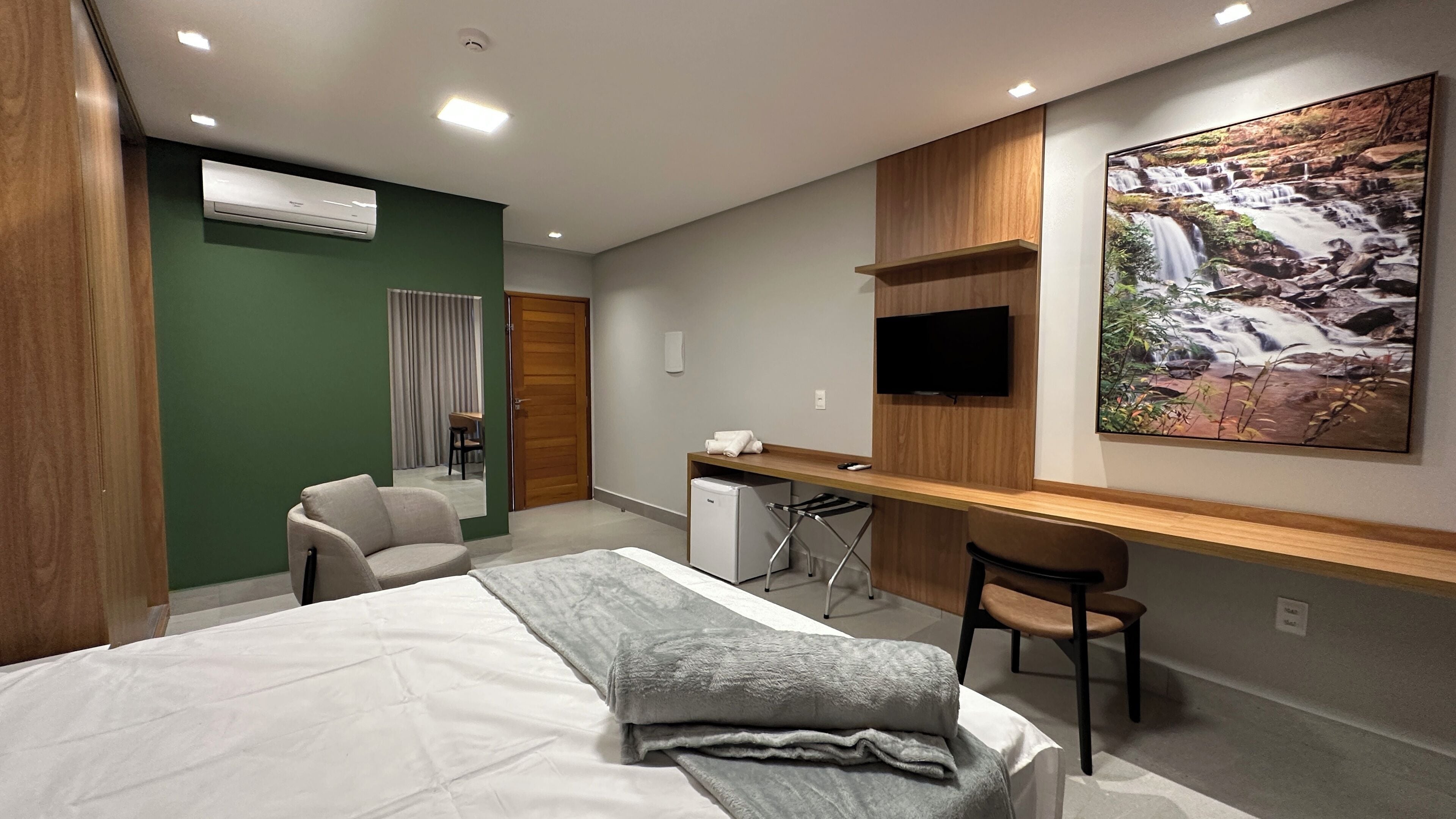 Habitación de hotel con cama, escritorio, silla, aire acondicionado, televisor y ventana grande con vistas a una cascada.
