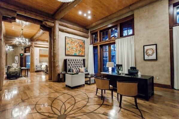 A spacious room with a large wooden ceiling, a tufted sofa, a desk with chairs, and a large window with curtains.