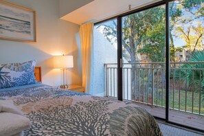 A bedroom with a bed, a nightstand, a lamp, a window with a view of trees and a balcony.