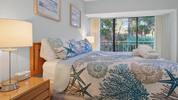 A bedroom with a bed, a nightstand, a lamp, a clock, and a window with a view of trees. - Sleeps 8!  2 Mins to Beach, 3 BD 3.5 BR- Sea Pines (Hilton Head Island)
