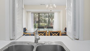 A kitchen with a double sink, a dining area with a glass table and chairs, and a view of a garden through a window with white shutters. - Sleeps 8!  2 Mins to Beach, 3 BD 3.5 BR- Sea Pines (Hilton Head Island)