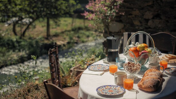 A breakfast table set with plates, glasses, a coffee pot, a fruit bowl, and a loaf of bread, overlooking a garden and a stone wall. - House of Artists (Xullán)