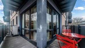 Un espace extérieur moderne doté d’une terrasse en bois, d’une porte vitrée donnant sur une pièce, ainsi que d’une table et d’une chaise en métal rouge.
