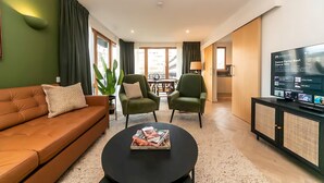 Un salon moderne comprenant un canapé, deux fauteuils, une table basse avec des magazines, un téléviseur et une grande fenêtre avec des rideaux.