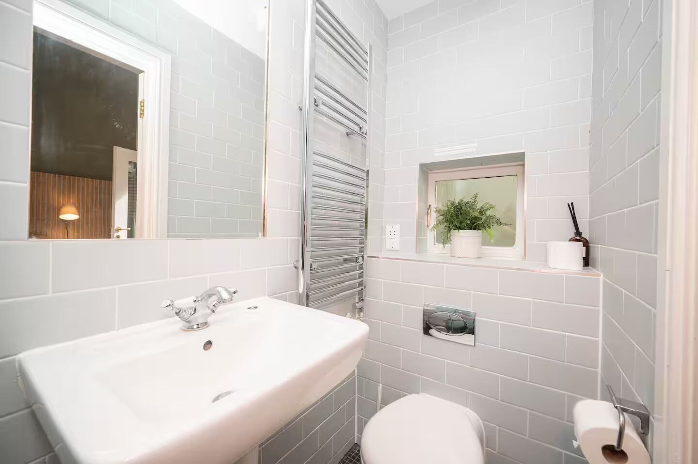 A modern bathroom with a white sink, a toilet, a window with a plant, and a towel rack.