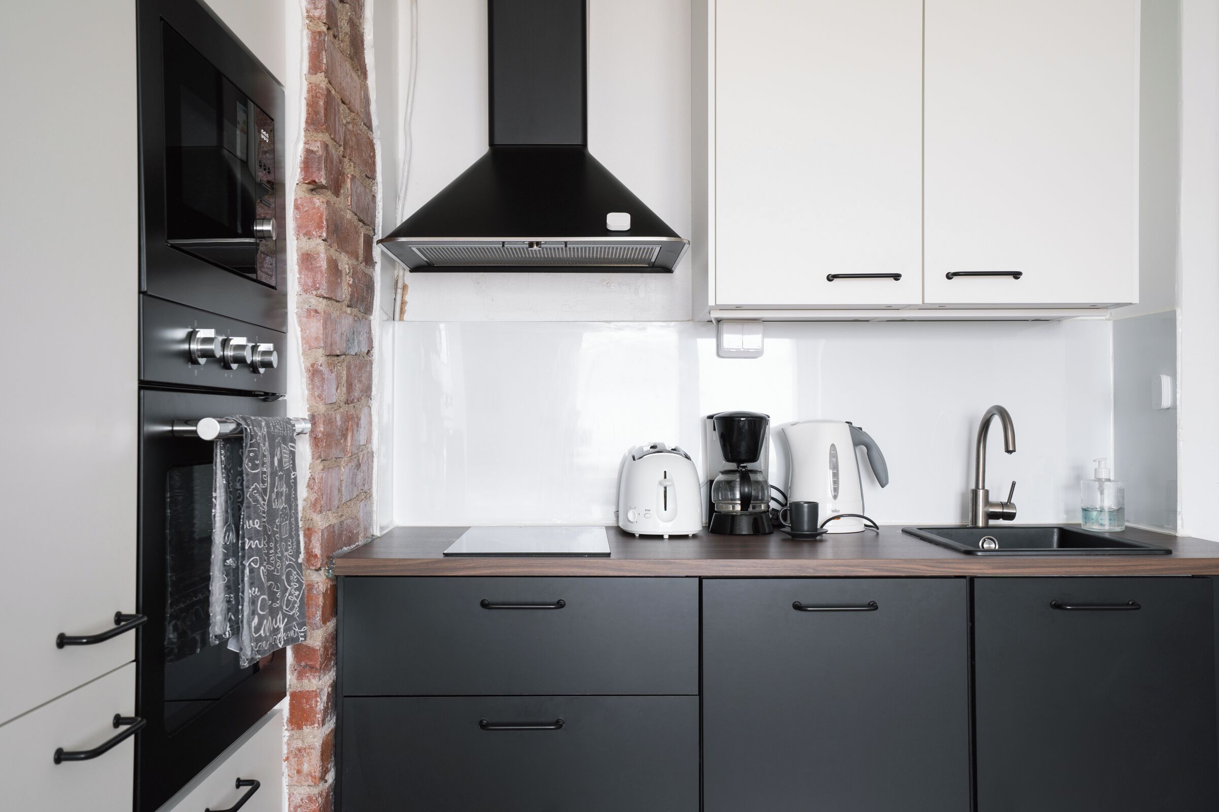 Una cocina moderna con gabinetes negros, una pared de acento de ladrillo, una campana extractora de acero inoxidable, una cafetera y un lavamanos.