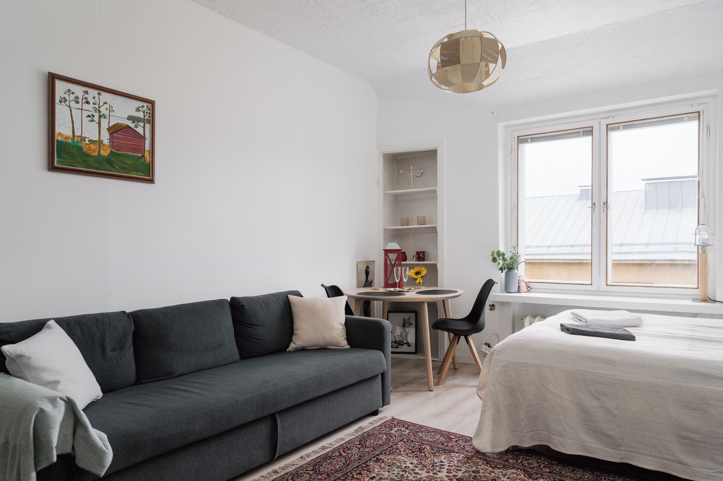 A modern living room with a grey sofa, a dining table with chairs, a bed with a white bedspread, a large window with blinds, and a painting on the wall.