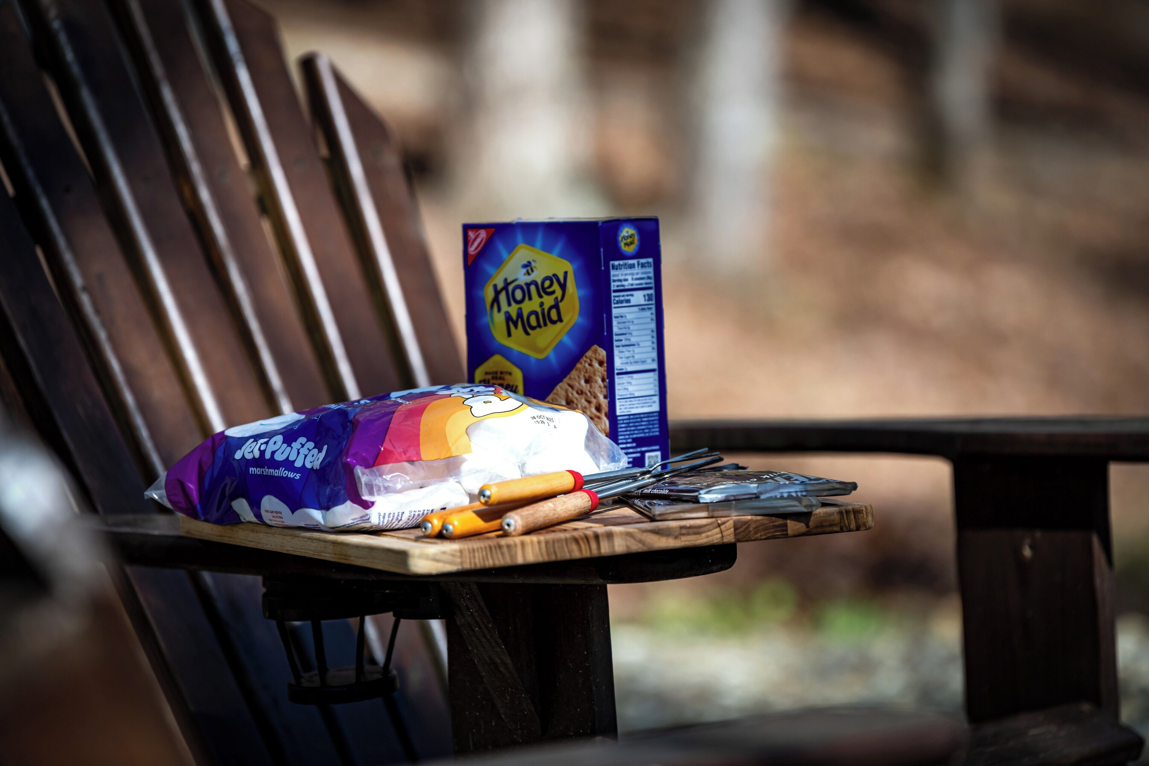A wooden bench with a package of Honey Maid cereal, a Jet-Puffed marshmallow, and a knife on it.