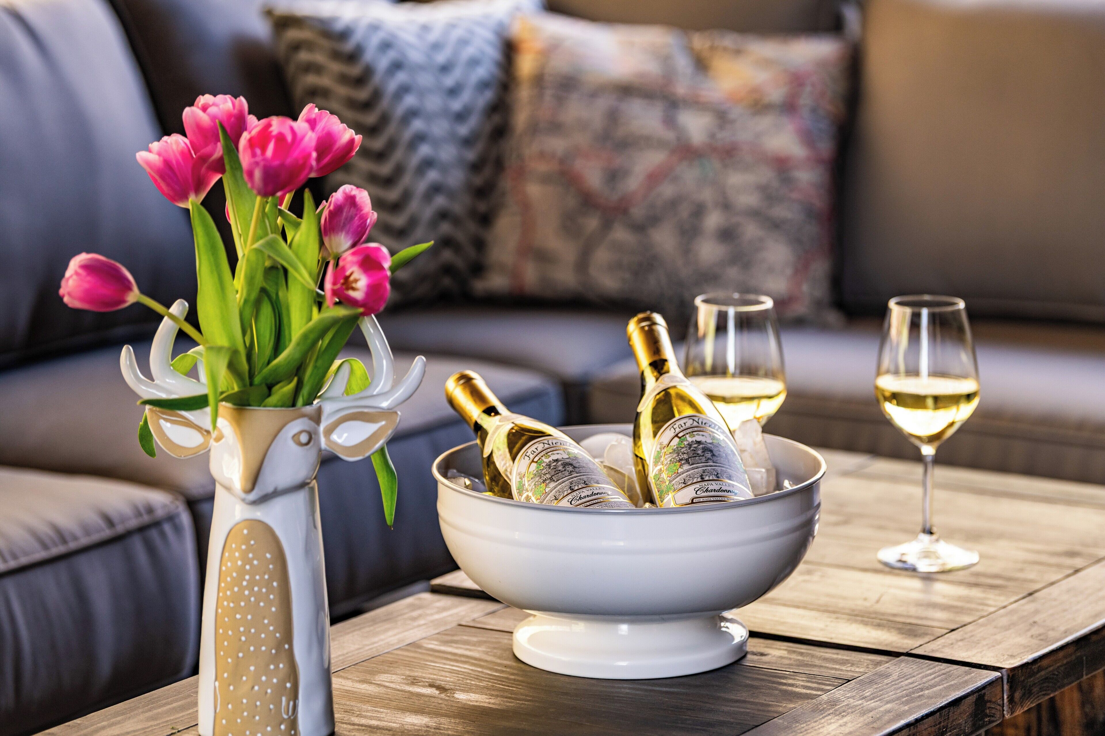 A wooden table with a white bowl containing champagne bottles and glasses, a white vase with pink tulips, and a grey sofa with patterned cushions in the background.