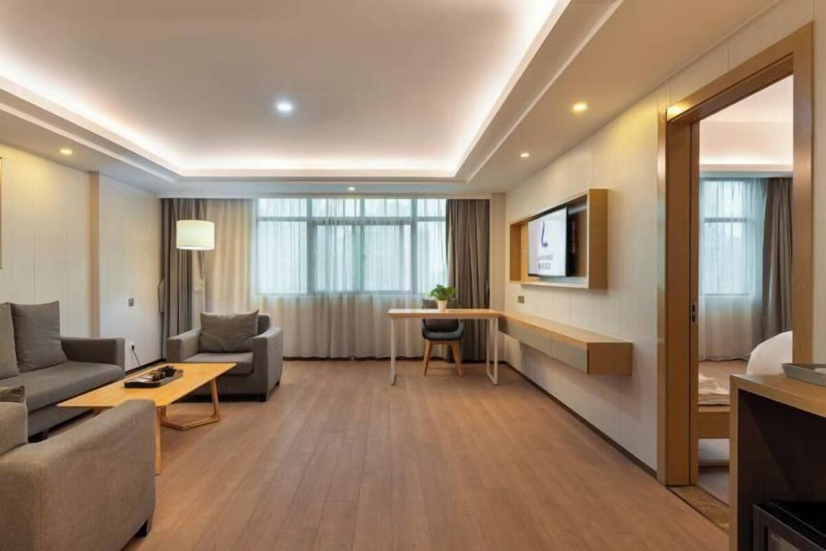 Ein modernes Hotelzimmer mit einer Sitzecke, einem kleinen Schreibtisch, einem an der Wand befestigten Fernseher und einem großen Fenster mit Vorhängen.